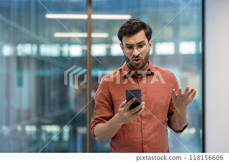 Man in office holding smartphone with confused expression, gesturing frustration. Business professional dealing with unexpected news or technical issues, showcasing emotions of surprise and disbelief 118156606