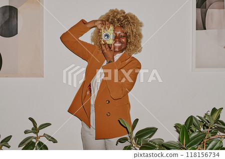 Smiling curly haired woman holding yellow camera and surrounded by ficuses while imitating photographer 118156734