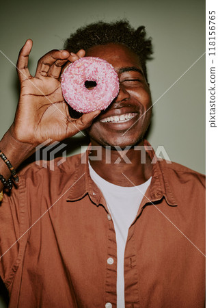 Young man holding pastel pink doughnut to hide his eye while posing for photo Young man holding pastel pink doughnut to hide his eye while posing for photo 118156765