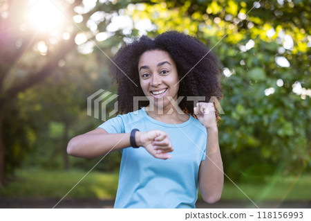 Woman in fitness clothing using smartwatch in park. Engaged in physical activity with enthusiasm and happiness. Enjoying nature and healthy lifestyle outdoors on a sunny day. 118156993