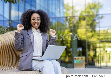 African American businesswoman sitting on bench with laptop, expressing excitement and success. Professional attire reflects confidence in urban environment, showcasing work culture and achievement African American businesswoman sitting on bench with laptop, expressing excitement and success. Professional attire reflects confidence in urban environment, showcasing work culture and achievement 118156998