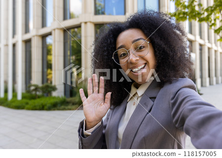 Confident African American business woman waving during phone video call. Standing in front of contemporary office building. Professional and friendly demeanor enhances business communication. 118157005