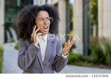 African American businesswoman experiencing frustration during phone call outside office. She expresses irritation. Urban corporate setting conveying stress, emotion, communication issues. 118157006