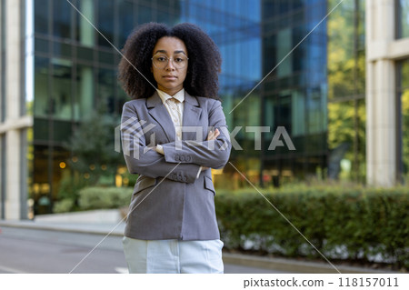 Confident African American businesswoman stands with arms crossed outside modern office building. Professional appearance reflects leadership, empowerment, success, and ambition in corporate setting 118157011