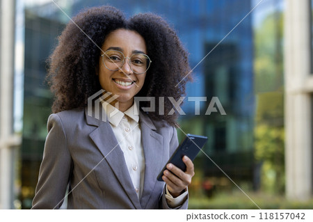 Smiling African American business woman in professional attire holding phone outside modern office building. Represents confidence, communication, technology in business environment Smiling African American business woman in professional attire holding phone outside modern office building. Represents confidence, communication, technology in business environment 118157042