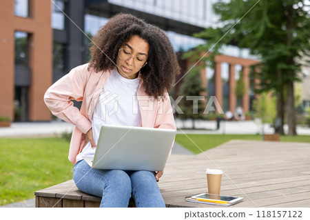 An African American woman is experiencing back pain while working on her laptop outdoors in a modern urban area. She is dressed casually with a coffee cup and tablet on the bench beside her. An African American woman is experiencing back pain while working on her laptop outdoors in a modern urban area. She is dressed casually with a coffee cup and tablet on the bench beside her. 118157122