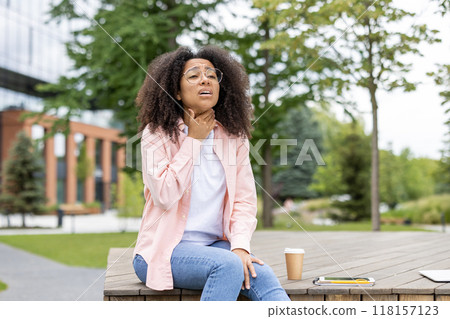 Woman sitting outside holding throat, expressing discomfort, possibly from soreness or pain. She is seated on wooden bench with phone and coffee cup visible. Urban park setting with greenery around. Woman sitting outside holding throat, expressing discomfort, possibly from soreness or pain. She is seated on wooden bench with phone and coffee cup visible. Urban park setting with greenery around. 118157123
