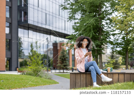 A young woman sits on a bench in a city park, using her smartphone. The modern building in the background adds a vibrant urban atmosphere. A young woman sits on a bench in a city park, using her smartphone. The modern building in the background adds a vibrant urban atmosphere. 118157133