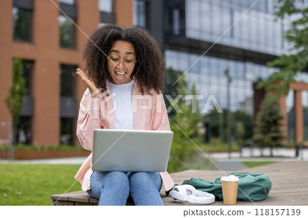African American student experiences excitement while working on laptop outdoors on college campus. Young adult studies with enthusiasm in modern educational setting, showcasing technology, learning African American student experiences excitement while working on laptop outdoors on college campus. Young adult studies with enthusiasm in modern educational setting, showcasing technology, learning 118157139