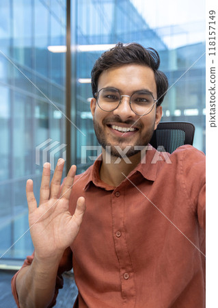 Cheerful young man with glasses waves during virtual meeting in modern office. Demonstrates concept of friendly communication, technology use, remote work, and professional interaction using phone in 118157149
