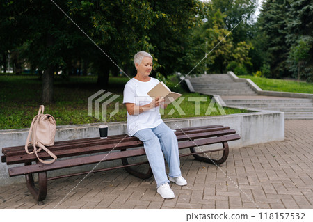 Full length portrait of older woman with short gray hair reading paper book in park, enjoying nature and tranquility, embodying relaxation and wisdom. Elderly female enjoying free time in retirement. Full length portrait of older woman with short gray hair reading paper book in park, enjoying nature and tranquility, embodying relaxation and wisdom. Elderly female enjoying free time in retirement. 118157532