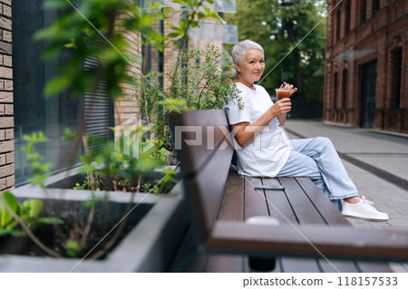 Side view of elegant elderly lady sitting on bench outdoors, savoring iced coffee and soaking in summer day. Happy European gray-haired older female takes moment to unwind and rejuvenate. Side view of elegant elderly lady sitting on bench outdoors, savoring iced coffee and soaking in summer day. Happy European gray-haired older female takes moment to unwind and rejuvenate. 118157533