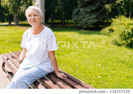 Serene senior woman with short white hair sitting on park bench, enjoying summer weather and looking away with happy expression. European gray-haired older female enjoying moment of relaxation 118157539