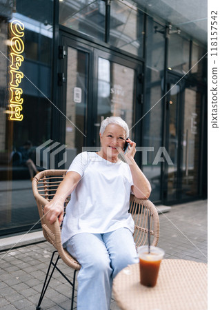Vertical portrait of cheerful senior female with short gray hair sipping iced coffee at street cafe, chatting on phone with smile, embracing joys of modern retirement, enjoying free time in pension. Vertical portrait of cheerful senior female with short gray hair sipping iced coffee at street cafe, chatting on phone with smile, embracing joys of modern retirement, enjoying free time in pension. 118157542
