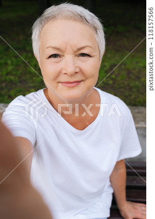 Vertical POV of attractive elderly woman with short grey hair capturing self-portrait while relaxing on park bench. Older female grandma user of modern tech looking on phone screen call grandkid Vertical POV of attractive elderly woman with short grey hair capturing self-portrait while relaxing on park bench. Older female grandma user of modern tech looking on phone screen call grandkid 118157566