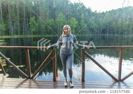 Young woman standing on a wooden dock against a serene lake surrounded by a dense forest. 118157580