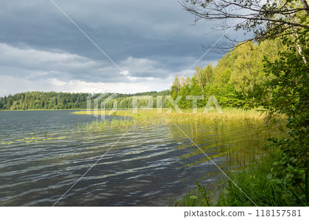 A serene lakeside scene with a lush, forested shoreline and a vibrant rainbow arching across the cloudy sky. 118157581