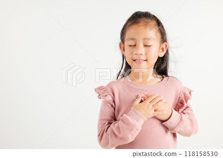 Happy young kid girl in casual clothes holding hands at chest close to heart meditation and smiling in studio short isolated on white background, kindergarten child believe faith and love gratitude 118158530