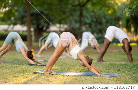 Multinational yoga enthusiasts with female coach perform Adho Mukha Svanasana in park Multinational yoga enthusiasts with female coach perform Adho Mukha Svanasana in park 118162717