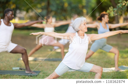 Elderly woman practices yoga pose Anjaneyasana with other people of different nationalities in summer park 118162874