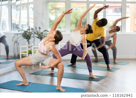 Young woman coach and students perform utkata konasana during training in studio Young woman coach and students perform utkata konasana during training in studio 118162941