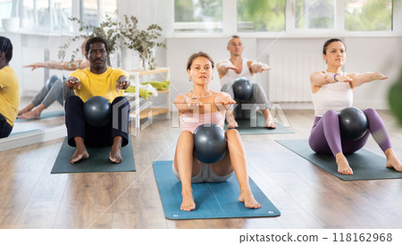 Young female Pilates instructor performing sit-ups with ball with group 118162968