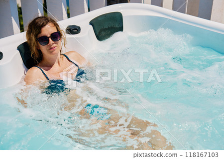 Woman wearing sunglasses and swimsuit relaxing in outdoor hot tub with bubbling water and enjoying sunny day Woman wearing sunglasses and swimsuit relaxing in outdoor hot tub with bubbling water and enjoying sunny day 118167103