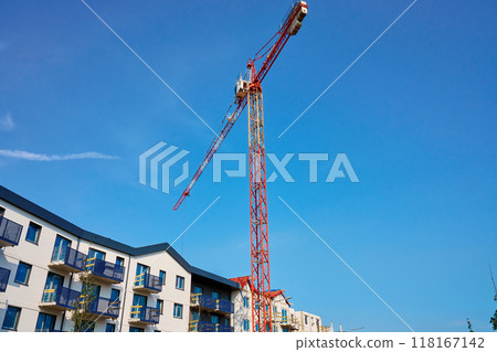 Red construction crane is building modern apartments in residential complex against blue sky. Construction of new housing and development of urban infrastructure. Real estate market Red construction crane is building modern apartments in residential complex against blue sky. Construction of new housing and development of urban infrastructure. Real estate market 118167142