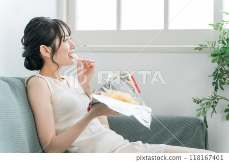 Young Asian woman eating potato chips on the sofa at home (binge eating, snacking, high calorie) 118167401