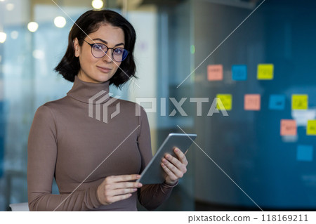 Confident businesswoman holding tablet in modern office. She stands next to colorful sticky notes. Ideal for concepts like productivity, technology, confidence, and office work. 118169211