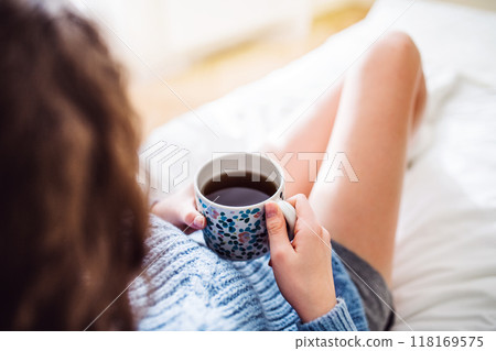 Morning cup of coffee. Beautiful young woman with curly hair sitting on bed in her room, drinking hot coffee. 118169575