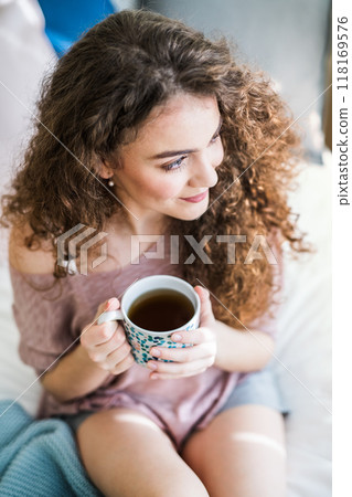 Morning cup of coffee. Beautiful young woman with curly hair sitting on bed in her room, drinking hot coffee. 118169576