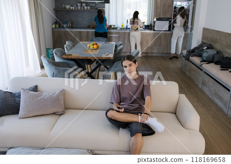 Cute teenage boy watch movie on tv in the living room, with three females preparing food in the kitchen in the background. The happy youngest brother always finds time to relax 118169658