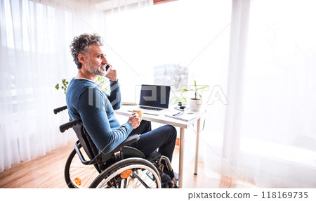 Handsome man in wheelchair working from home office, making phone call. Telecommuting and home office. Handsome man in wheelchair working from home office, making phone call. Telecommuting and home office. 118169735