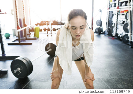 Young fitness girl with sweat on face feeling exhausted after hard workout in gym, holding knees, breathing. Young fitness girl with sweat on face feeling exhausted after hard workout in gym, holding knees, breathing. 118169749