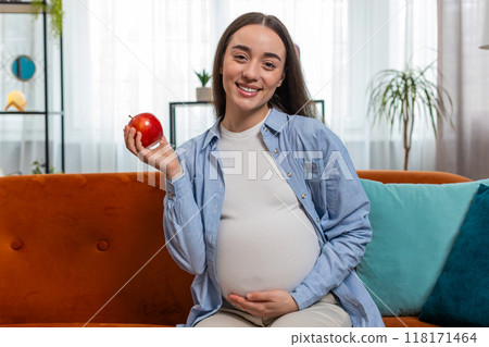 Happy pregnant woman eating fresh red apple fruit sitting on sofa healthy vitamin food motherhood 118171464
