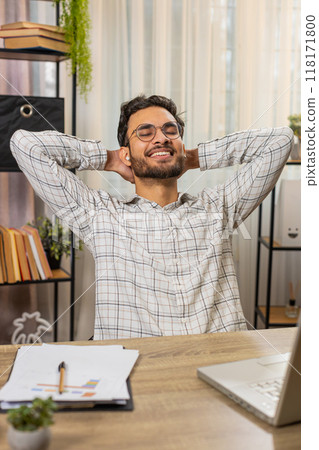 Indian businessman working on laptop putting on earbuds listening music taking a break relaxing 118171800