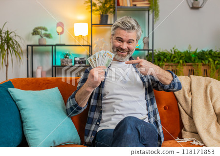 Amazed middle-aged man showing pointing money dollar cash bills clenching fist thumbs up at home 118171853