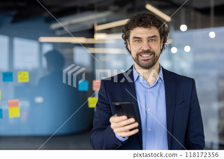 Confident professional man holding phone, smiling in modern office. Dressed in blue suit, communicates digital success and connectivity. Ideal for concepts like business 118172356