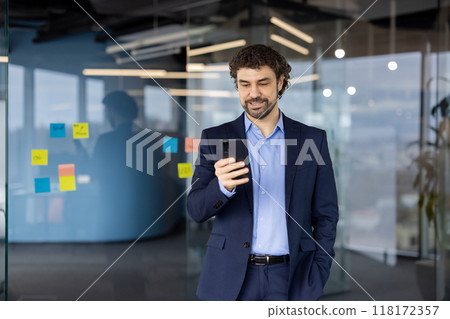 Businessman in a blue suit uses smartphone in modern office setting. Glass walls filled with colorful sticky notes highlight focus on innovation and communication. 118172357