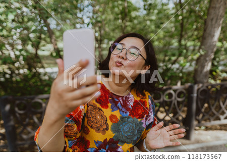 Smiling freelancer wearing a colorful blouse taking a selfie outdoors with her smartphone Business casual professional taking a selfie on her cell phone while enjoying nature 118173567