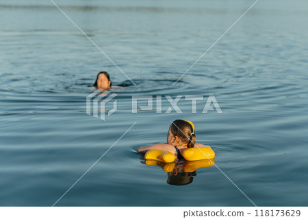 Young girl floating with a yellow duck float at the lake. Her mother is watching over her, ensuring a safe and enjoyable swim Young girl floating with a yellow duck float at the lake. Her mother is watching over her, ensuring a safe and enjoyable swim 118173629