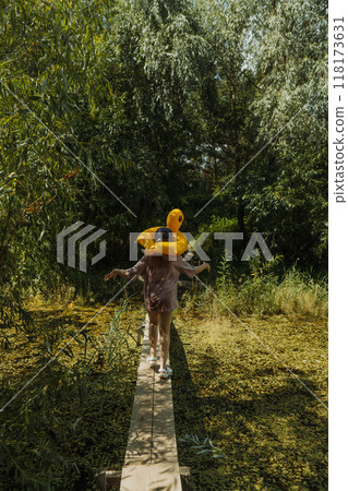 A girl is walking on a wooden bridge through a pond with a yellow duck float on her head. The photo is taken from behind, showing no face A girl is walking on a wooden bridge through a pond with a yellow duck float on her head. The photo is taken from behind, showing no face 118173631