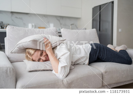 A woman appears stressed, lying on a couch with a pillow overhead in a modern home environment A woman appears stressed, lying on a couch with a pillow overhead in a modern home environment 118173931