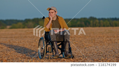 A man in a wheelchair sits in an empty field, resting his head on his hand, looking thoughtful. 118174199