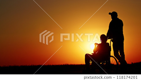 Silhouette of a person in a wheelchair being pushed by another person at sunset. Silhouette of a person in a wheelchair being pushed by another person at sunset. 118174201