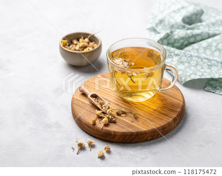 Chamomile herbal tea in a glass cup on a wooden board on a light background with dry flowers. Chamomile herbal tea in a glass cup on a wooden board on a light background with dry flowers. 118175472
