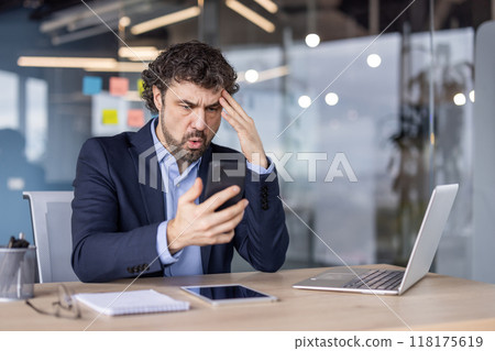 Businessman in suit experiencing frustration while looking at phone in modern office. Emotional reaction to message or email, working with laptop and tablet at desk. 118175619