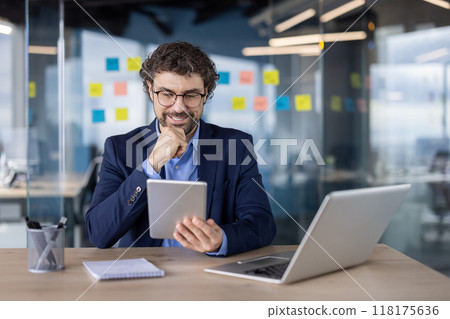 Confident businessman in suit using a digital tablet at a modern office desk with laptop and notepad. Bright workspace and innovative atmosphere. 118175636