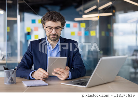 Business professional using tablet and laptop at desk in modern office. Man wearing glasses and suit, working efficiently. Focus is on technology and productivity. Business professional using tablet and laptop at desk in modern office. Man wearing glasses and suit, working efficiently. Focus is on technology and productivity. 118175639